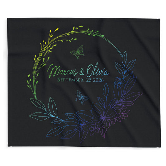 A black square fabric item, likely a blanket or throw, featuring a personalized floral wreath design with the names 'Marcus & Olivia' and the date 'September 25 2026' in a green-to-blue-to-purple gradient.