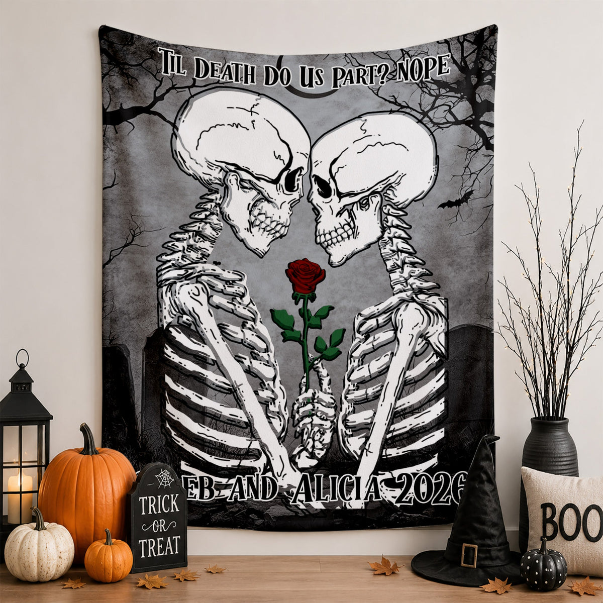 A Halloween-themed wall tapestry featuring two facing skeletons holding a single red rose, displayed on a wall above a decorated tabletop with pumpkins, a lantern, a witch hat, and a decorative pillow.