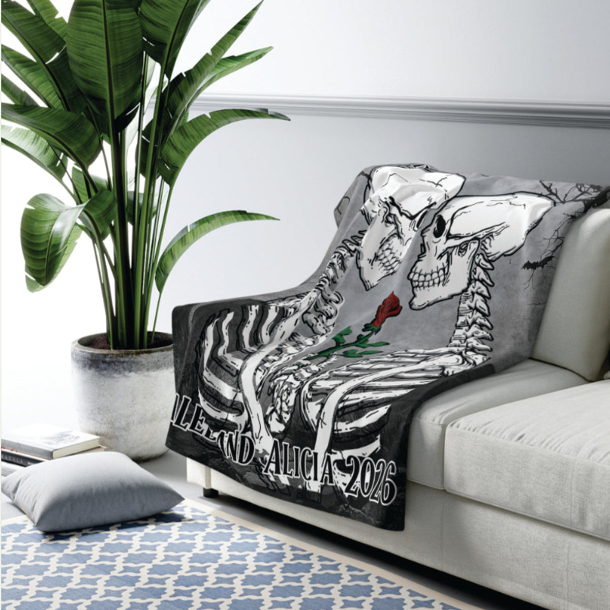 A throw blanket draped over a light gray sofa in a living room setting, featuring a black-and-white skeleton illustration with a small red rose and the text 'MILE AND ALICIA 2026'. A large green potted plant and a blue patterned rug are visible in the background.
