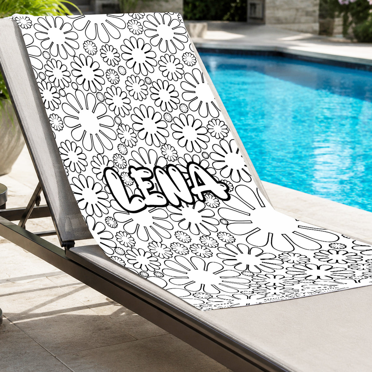 A white beach towel with a black daisy pattern and the name 'LENA' in a bold outlined font, draped over a beige lounge chair next to a swimming pool.