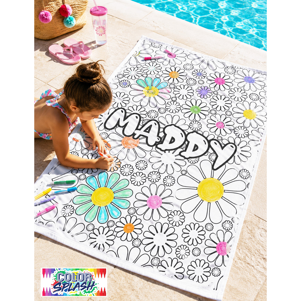A child coloring on a large white outdoor mat with black floral outlines and the word “MADDY” in large letters. The mat is placed on a patio beside a swimming pool, with colored markers and a straw hat nearby.