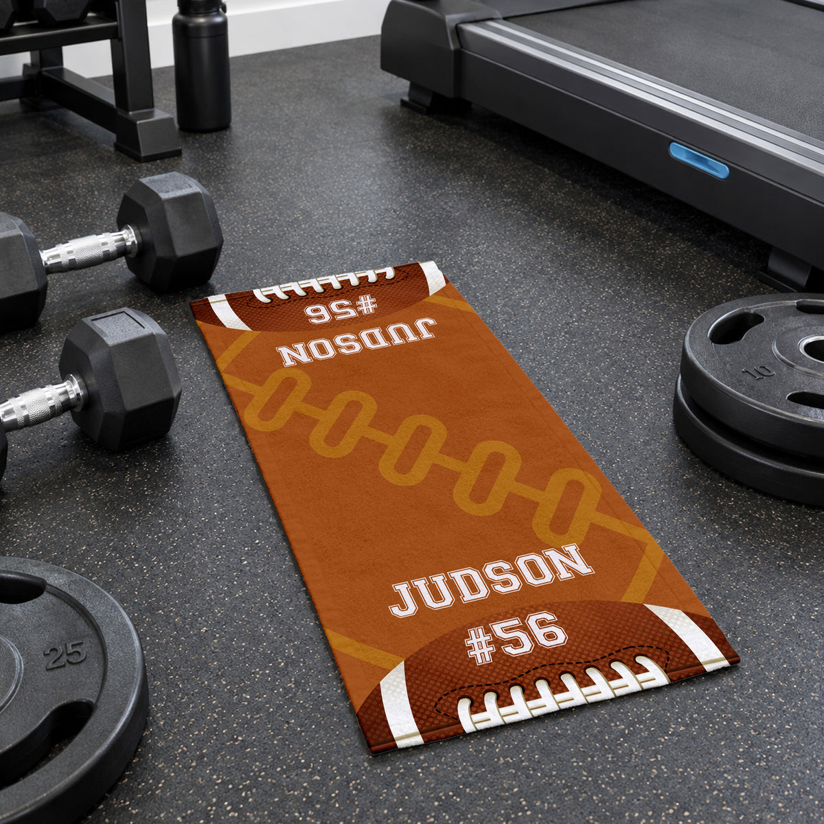 A custom football-themed gym mat lies on a dark gym floor, surrounded by dumbbells, weight plates, and a treadmill. The mat features a brown football design with white laces, white text reading 'JUDSON #56', and yellow chain graphics.