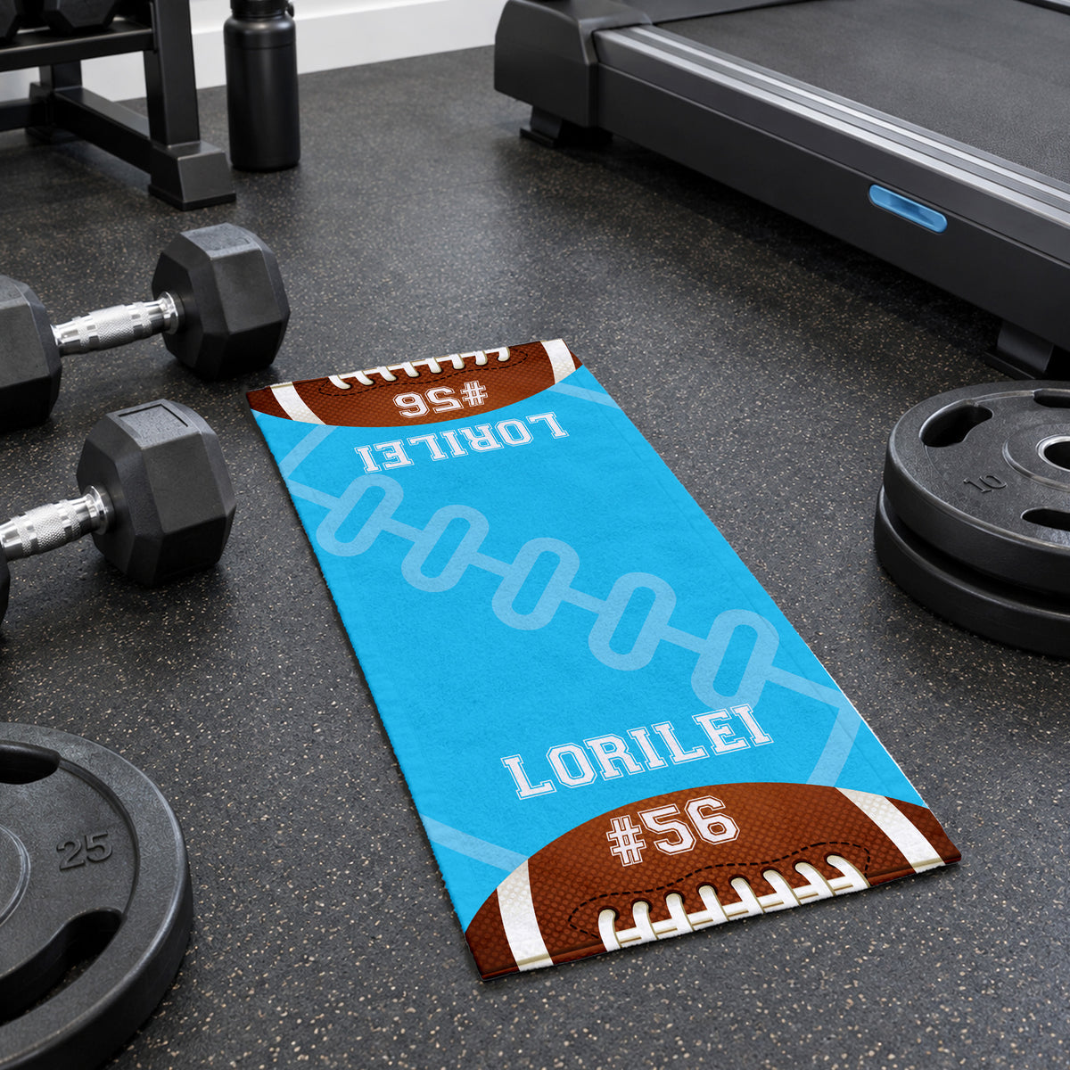 A custom football-themed runner mat on a dark gym floor, surrounded by dumbbells, a treadmill, and weight plates. The mat features a bright blue background with white football laces, brown football ends, and white text reading 'LORILEI' and '#56'.