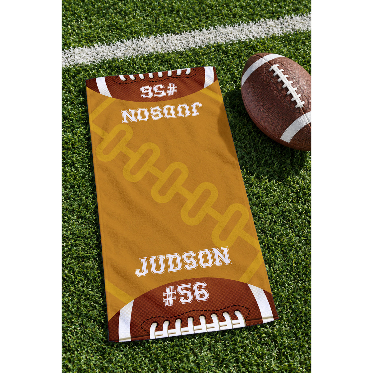 A custom football-themed towel laid out on green turf next to a brown football. The towel is gold with white and brown details, featuring the name 'JUDSON' and number '#56' printed on it.