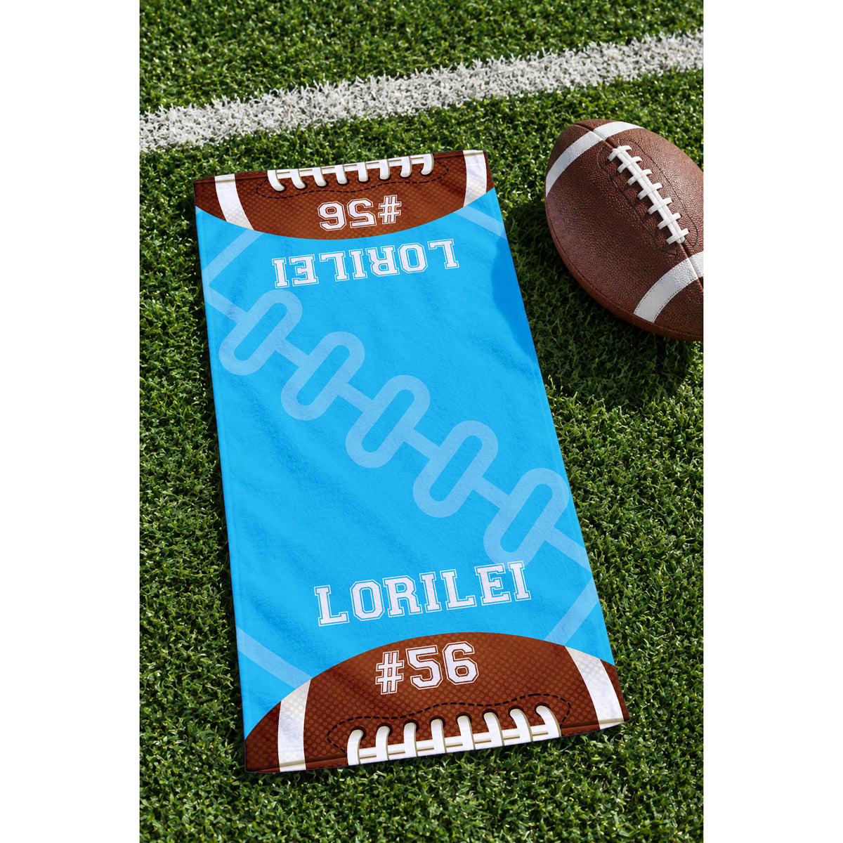 A custom football-themed beach towel laid out on green turf next to a brown football. The towel features a bright blue center with white football laces and the name 'LORILEI' and number '#56' printed in white, with brown football ends at the top and bottom.
