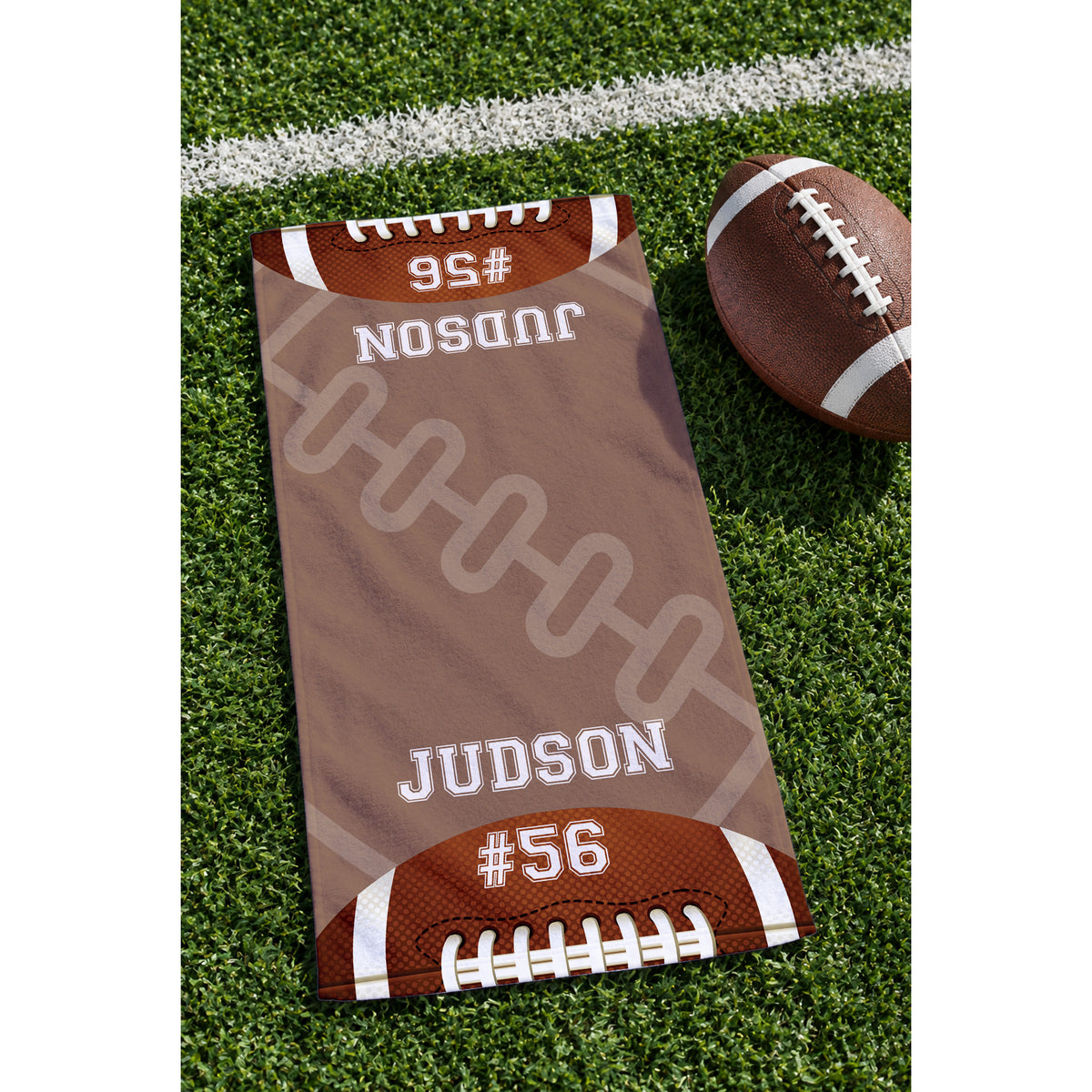 A custom football-themed towel laid out on a green turf field next to a brown leather football. The towel features a brown background with white laces graphic, white text reading 'JUDSON' and '#56', and brown football ends with white stripes.