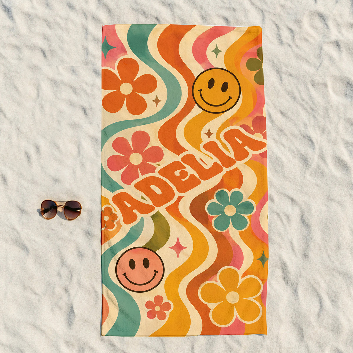 A rectangular beach towel with a retro 1960s-style wavy pattern in orange, teal, pink, and cream, featuring smiley faces and the name 'ADELIA'. The towel is laid flat on white sand next to a pair of sunglasses.
