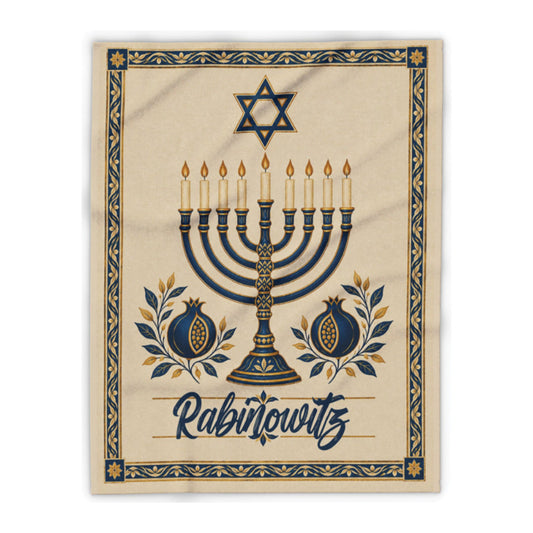 A square beige textile print or blanket featuring a decorative Hanukkah design with a menorah, Star of David, pomegranates, and the word 'Rabinowitz' in script, shown on a white background.