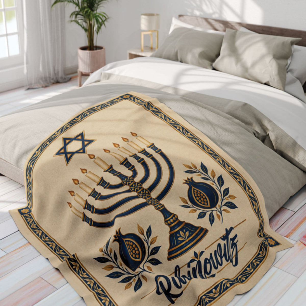 A beige blanket with a Hanukkah design, draped over a bed in a bright, modern bedroom. The blanket features a navy blue menorah, Star of David, pomegranate, and the word 'Purim' in navy script.
