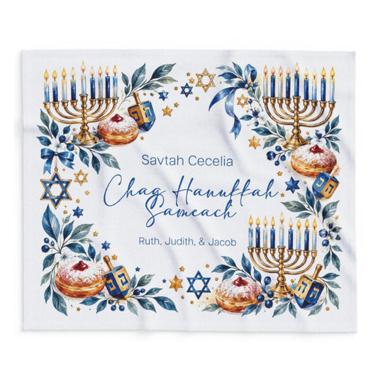 A square white fabric item, likely a towel or blanket, printed with a festive Hanukkah border design. The design features watercolor illustrations of gold and blue menorahs, dreidels, Star of David emblems, blue leaves, and pastries, surrounding personalized text that reads 'Savtah Cecelia Chag Hanukkah Sameach Ruth, Judith, & Jacob'.