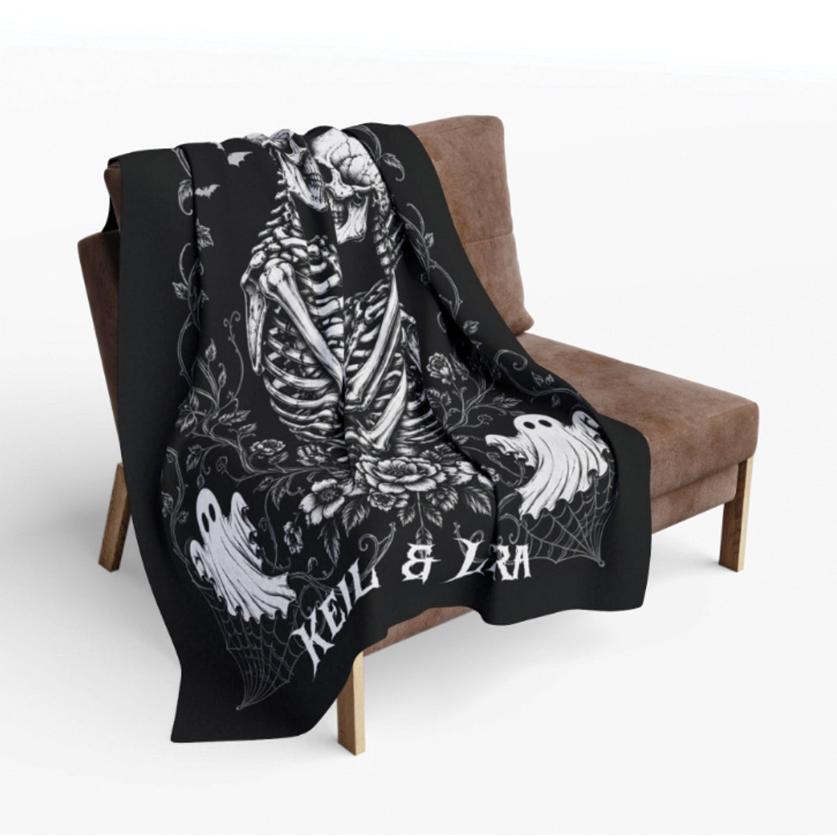 A black throw blanket draped over a brown armchair, featuring a large white skeleton illustration, floral details, and the text 'KEEL & LRA'.