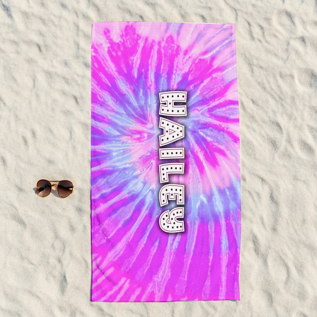 A rectangular beach towel with a vibrant pink, purple, and blue tie-dye spiral pattern, featuring the word 'HARLEY' printed vertically in white block letters with black dots. The towel is laid flat on white sand next to a pair of sunglasses.