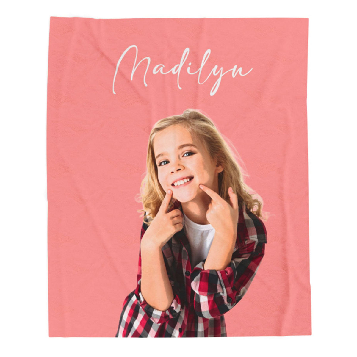 A personalized blanket featuring a photo of a smiling young girl with blonde hair, wearing a red and black plaid flannel shirt over a white t-shirt. The blanket has a solid coral pink background with the name 'Madilyn' written in white cursive script at the top.