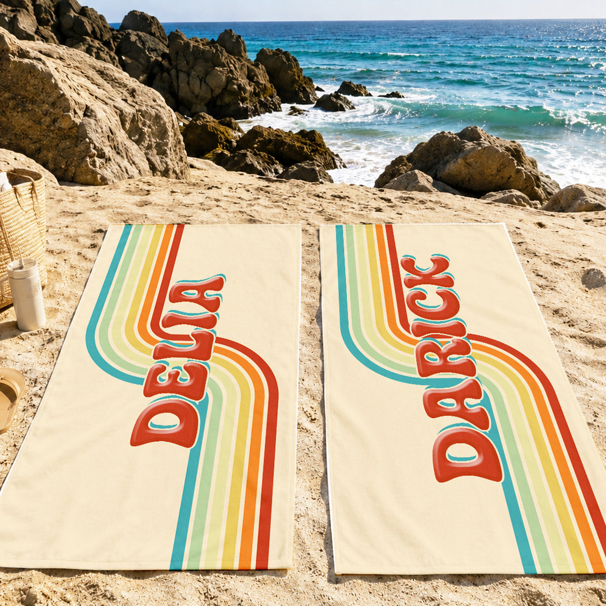 Two cream-colored beach towels laid out on sandy beach near the ocean, featuring retro rainbow stripes and the names 'DELIA' and 'DARICE' printed vertically in red.