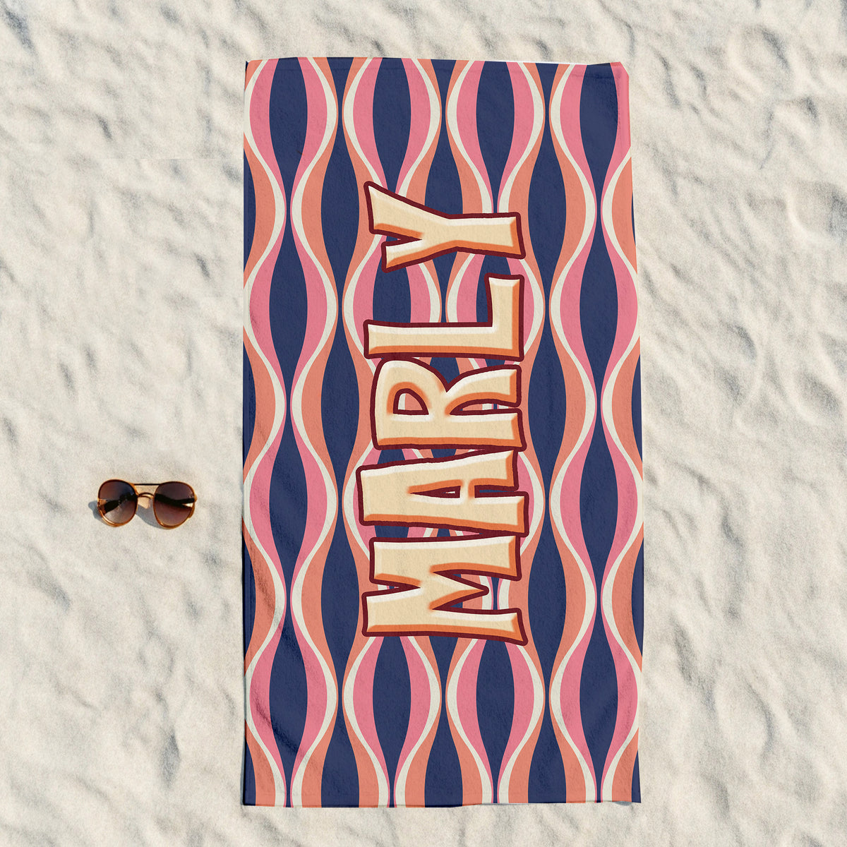 A rectangular beach towel laid flat on white sand, featuring a retro wavy pattern in navy, pink, peach, and cream, with the name 'MARLY' printed vertically in large 3D-style letters. A pair of sunglasses rests to the left of the towel.