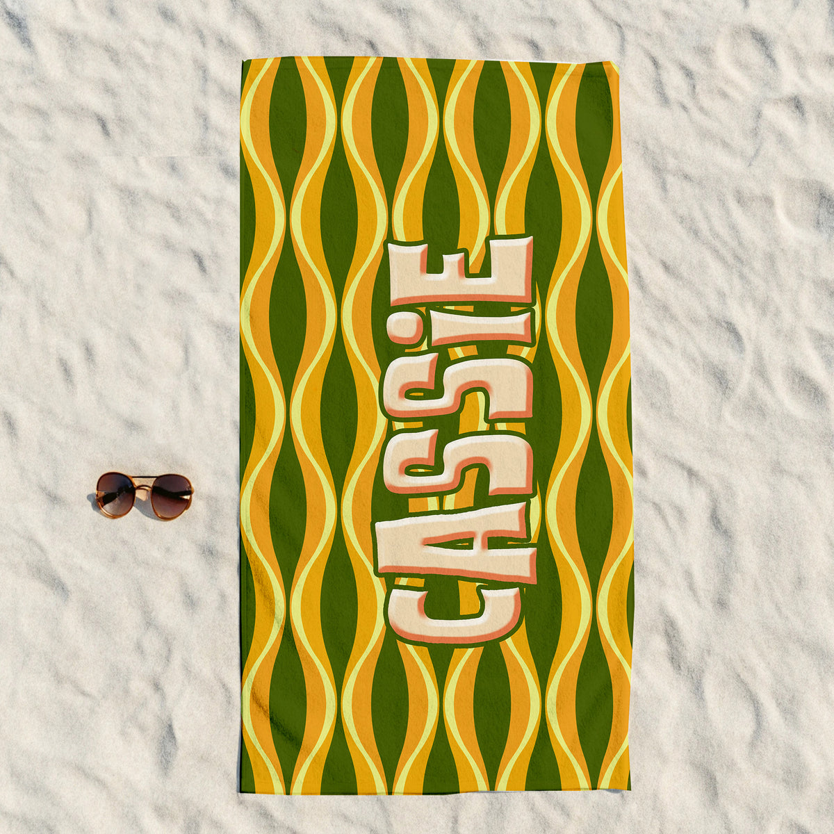 A rectangular beach towel laid flat on white sand, featuring a retro wavy pattern in olive green and mustard yellow with the name 'CASSIE' printed vertically in large cream letters with a pink outline. A pair of sunglasses rests to the left of the towel.