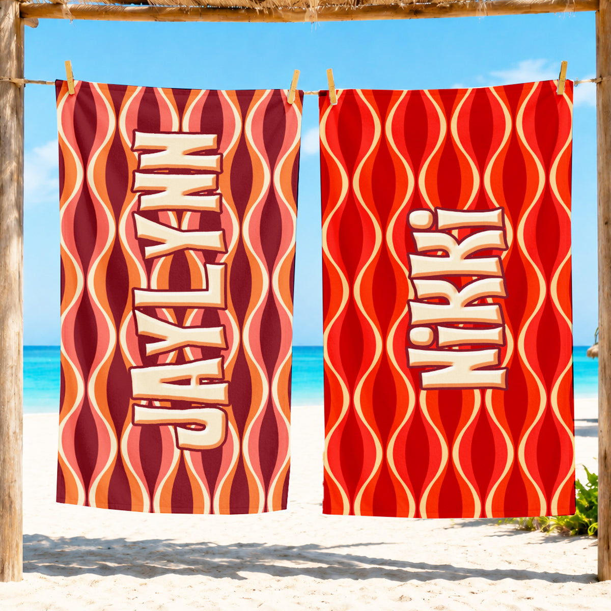 Two beach towels hanging from a wooden frame at the beach, featuring a retro wavy pattern in red, orange, and cream with the word 'JAXLXN' printed vertically.