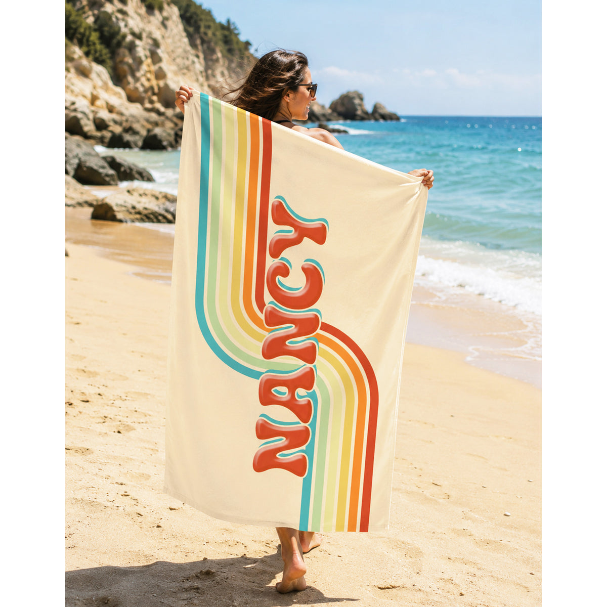 A woman on a sandy beach holds up a large cream-colored beach towel featuring a retro rainbow design and the word 'NANCY' in large red letters.