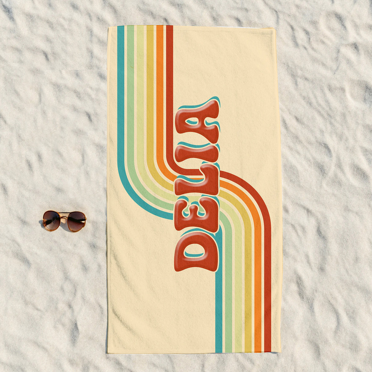 A rectangular beach towel laid flat on white sand, featuring retro vertical rainbow stripes and the word 'DELTA' in large red-orange letters outlined in teal. Small sunglasses sit to the left of the towel.