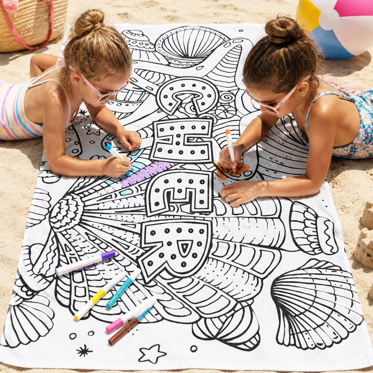 May include: A white rectangular beach towel featuring a black line art design of various seashells and the word BEACH printed in large block letters. The towel is scattered with several colorful markers used for coloring the printed illustrations.
