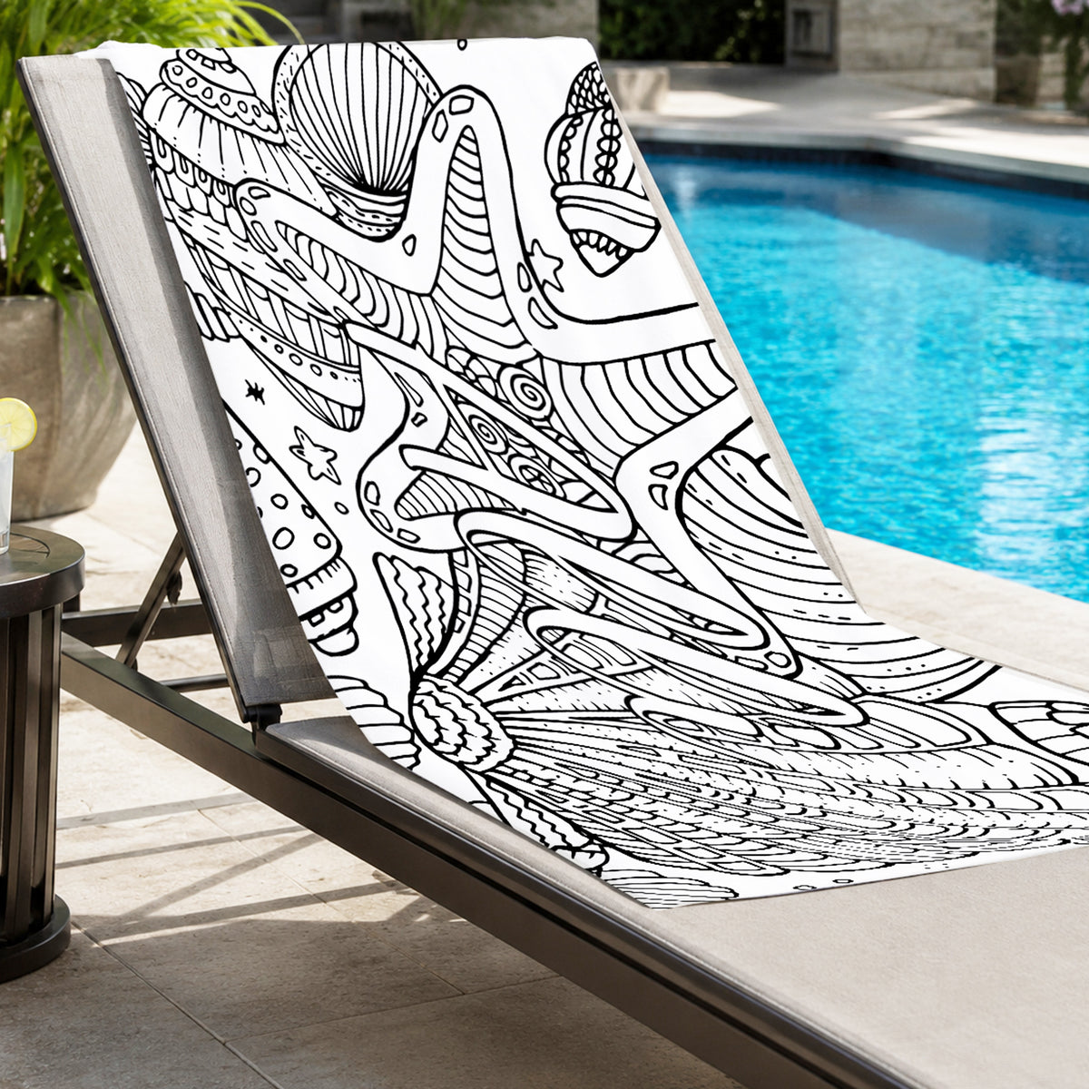 A black and white seashell and starfish patterned blanket is draped over a beige outdoor lounge chair next to a swimming pool.