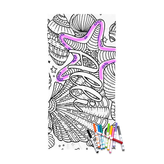 May include: A white rectangular beach towel featuring a black line art design of various seashells and the word BEACH printed in large block letters. The towel is scattered with several colorful markers used for coloring the printed illustrations.