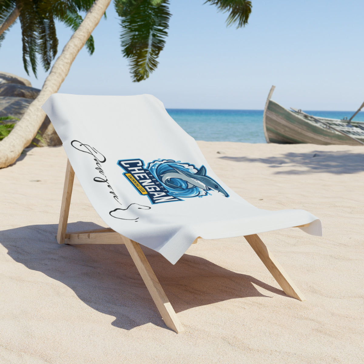 A white beach towel with a blue and yellow 'Chee Gang' graphic and black script lettering, draped over a wooden lounge chair on a sandy beach with palm trees and the ocean in the background.