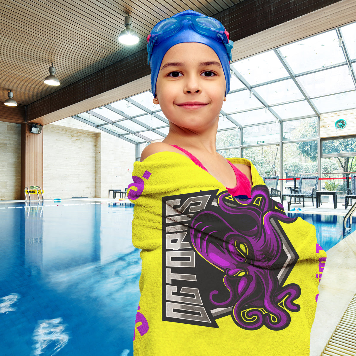A young girl wearing a blue swim cap and goggles stands in an indoor swimming pool, wrapped in a bright yellow towel featuring a purple octopus graphic and the text 'Octopus'.