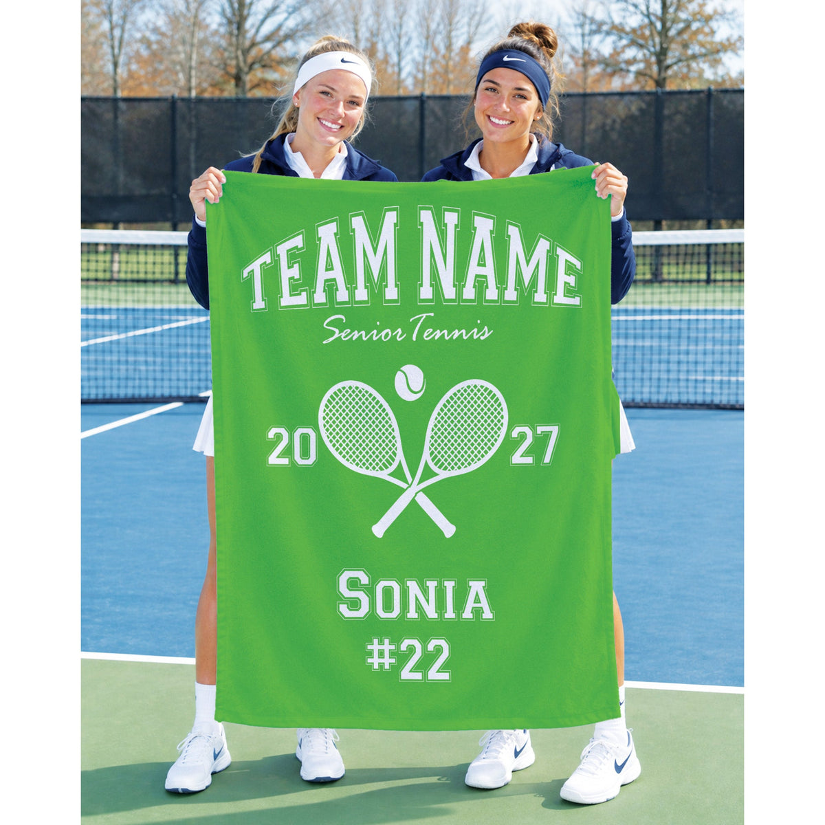 Two women standing on a tennis court holding a bright green personalized banner that reads 'TEAM NAME Senior Tennis' with crossed tennis rackets, the numbers 2027, and 'SONIA #22'.