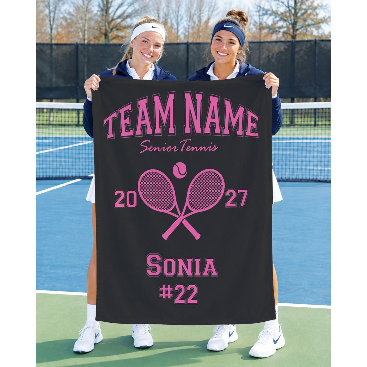 Two women standing on a tennis court holding a black personalized banner with pink text reading 'TEAM NAME Senior Tennis' and 'SONIA #22,' plus pink crossed tennis rackets and numbers 2027.