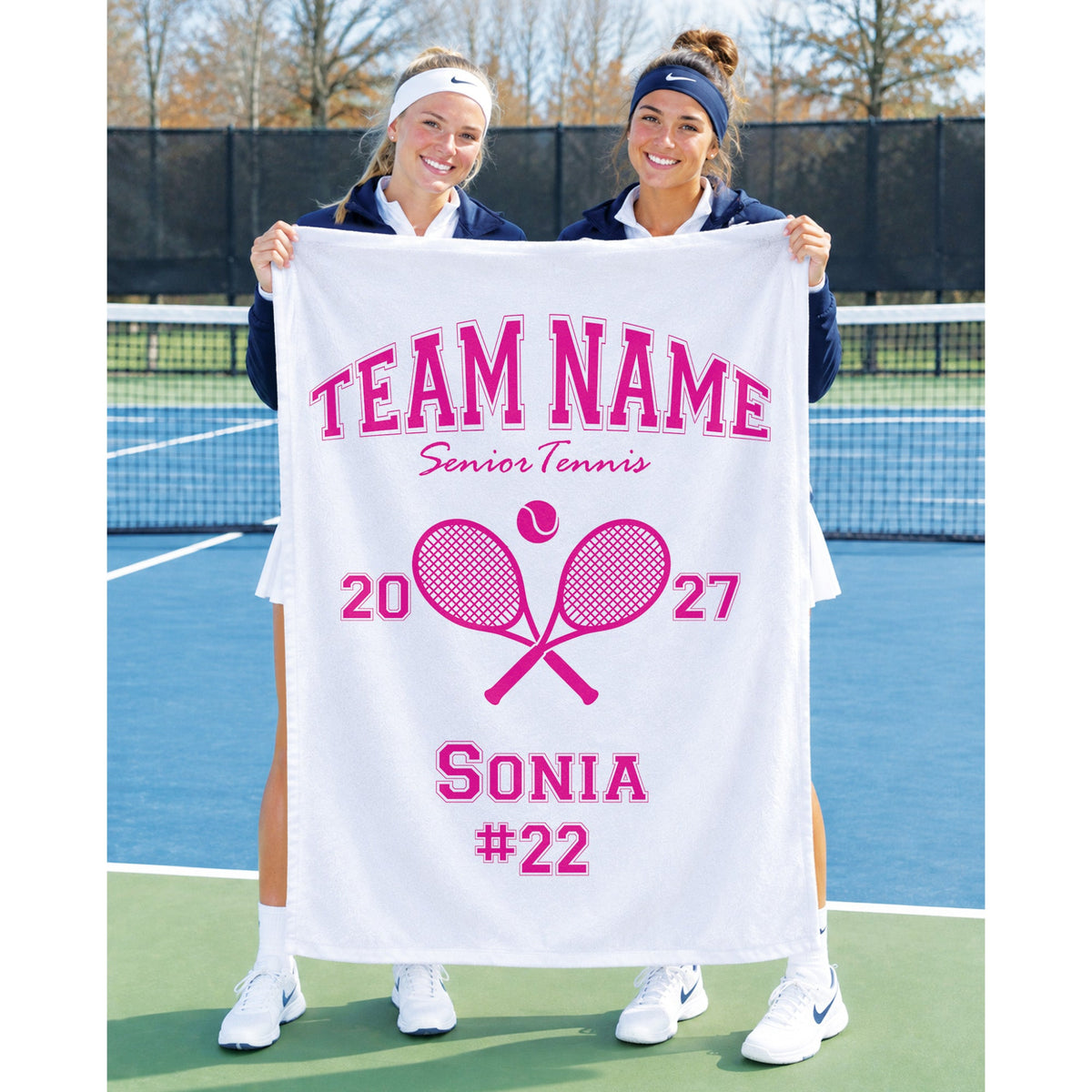 Two women standing on a tennis court holding up a large white personalized towel with pink text and graphics reading 'TEAM NAME Senior Tennis 2027 Sonia #22' and crossed tennis rackets.