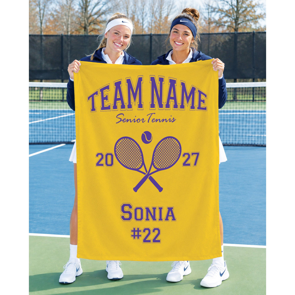 Two women standing on a tennis court holding up a large personalized yellow banner that reads 'TEAM NAME Senior Tennis 2027 SONIA #22' with crossed tennis rackets in the center.