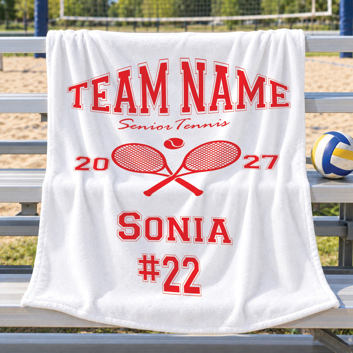 A white fleece blanket draped over a metal bench at a sandy beach volleyball court, printed with red text reading 'TEAM NAME Senior Tennis 2027 SONIA #22' and crossed tennis rackets.