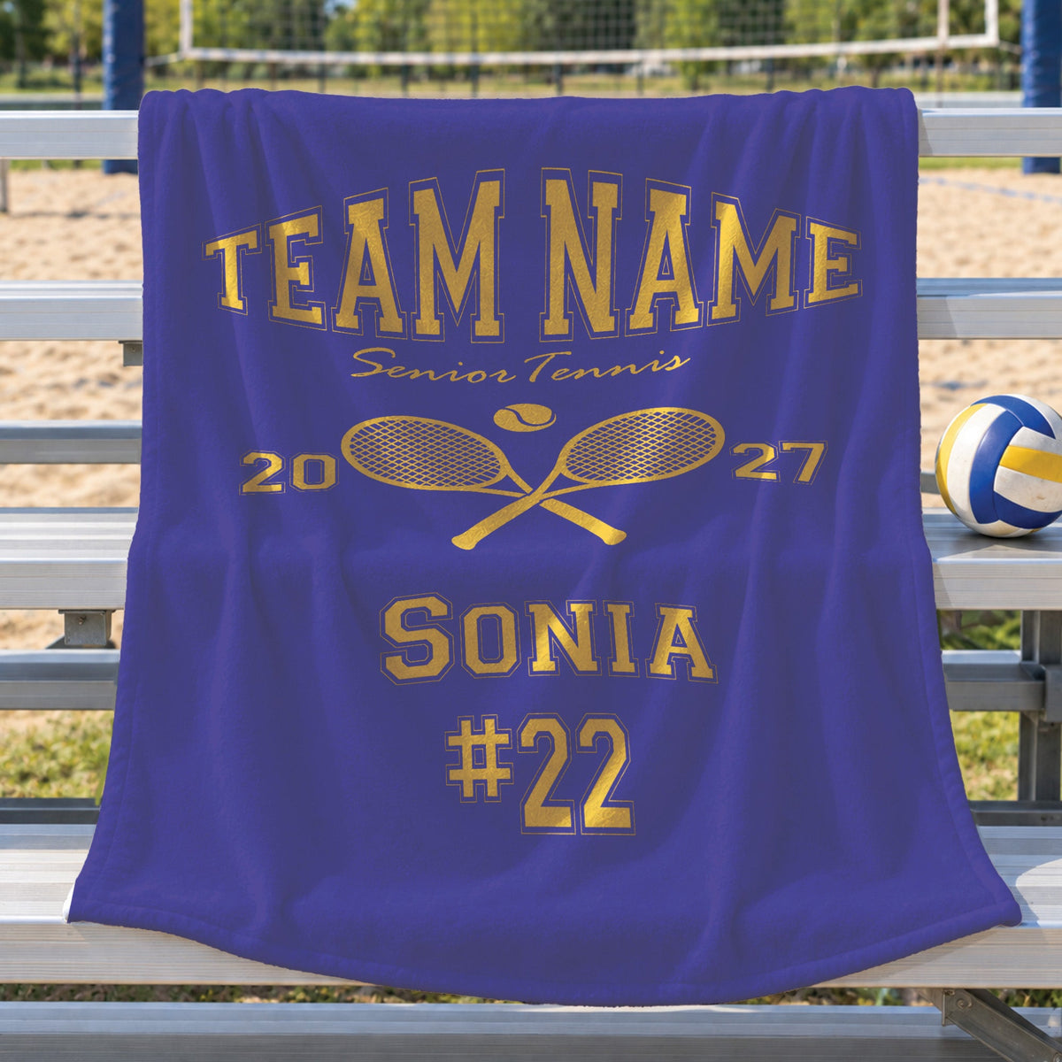 A royal blue blanket draped over a metal bench at a volleyball court, printed with gold text reading 'TEAM NAME Senior Tennis 2027 SONIA #22' and crossed tennis rackets.