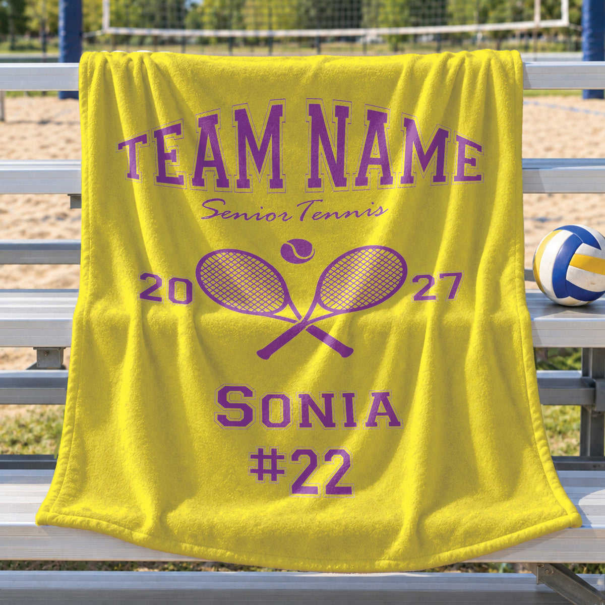 A bright yellow personalized blanket draped over a metal bench at a tennis court, featuring purple text that reads 'TEAM NAME Senior Tennis 2027 SONIA #22' with crossed tennis rackets and a tennis ball graphic. A volleyball sits on the bench beside it.