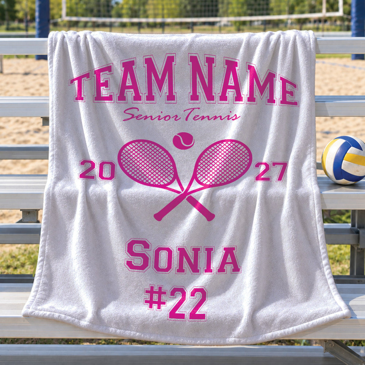 A white terry cloth towel draped over a metal tennis court bench, featuring a bright pink custom print with the text 'TEAM NAME Senior Tennis', crossed tennis rackets, '2027', and 'SONIA #22'. A volleyball sits on the bench next to it, with a sandy court and net visible in the blurred background.