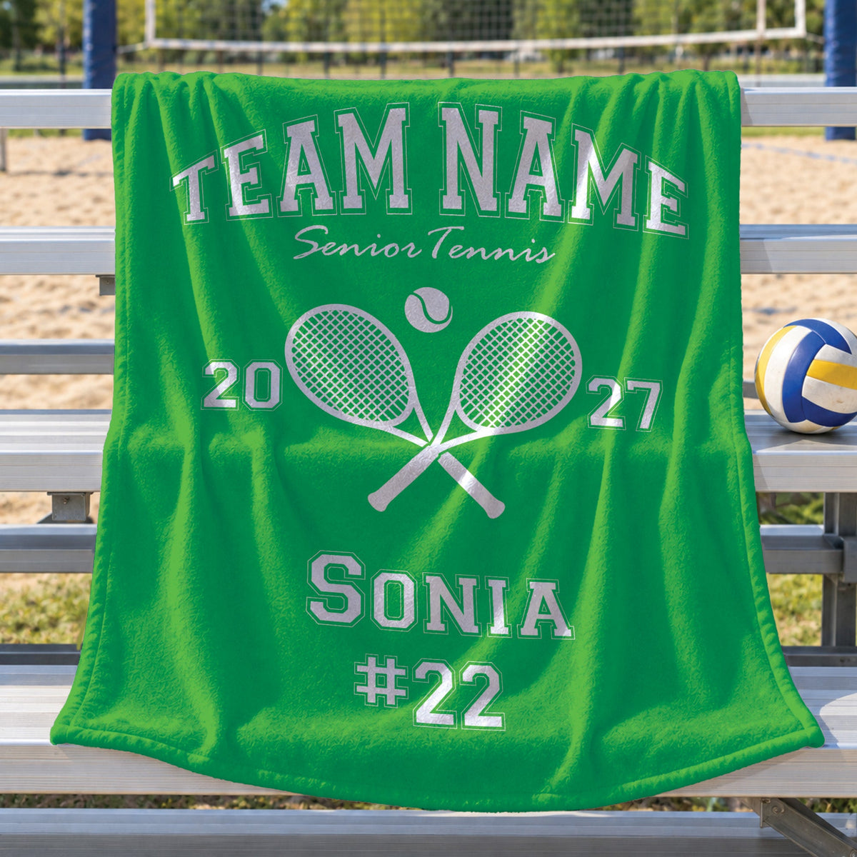 A bright green custom blanket draped over a metal tennis court bench, featuring white text that reads 'TEAM NAME Senior Tennis' with crossed rackets, the number '2027', and 'SONIA #22'. A volleyball sits on the bench next to it, with the sandy court and net visible in the blurred background.