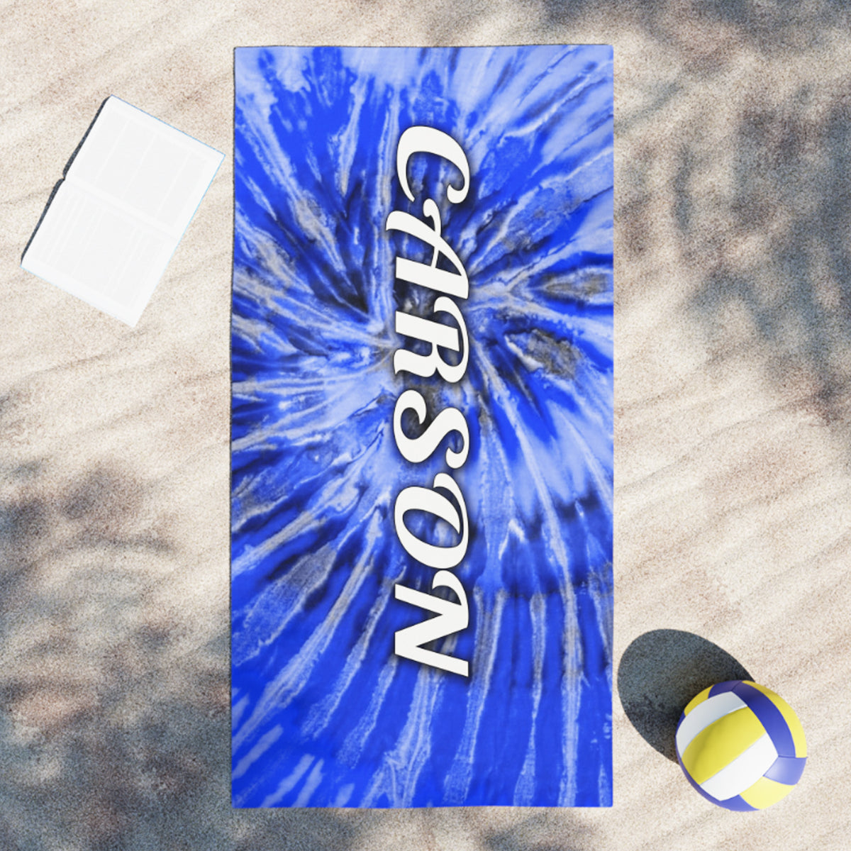 A rectangular beach towel laid flat on sand, featuring a blue and white tie-dye burst pattern with the name 'CARSON' printed vertically in white. A white folded towel, a yellow and blue volleyball, and a dark shadow are visible around it.