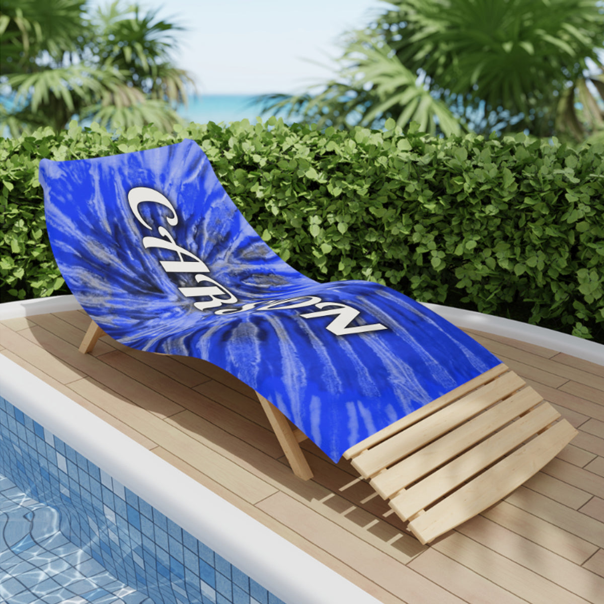 A blue and white tie-dye lounge chair blanket with the word 'CHILL' printed in white, displayed on a wooden poolside chaise lounge in front of a hedge and ocean view.