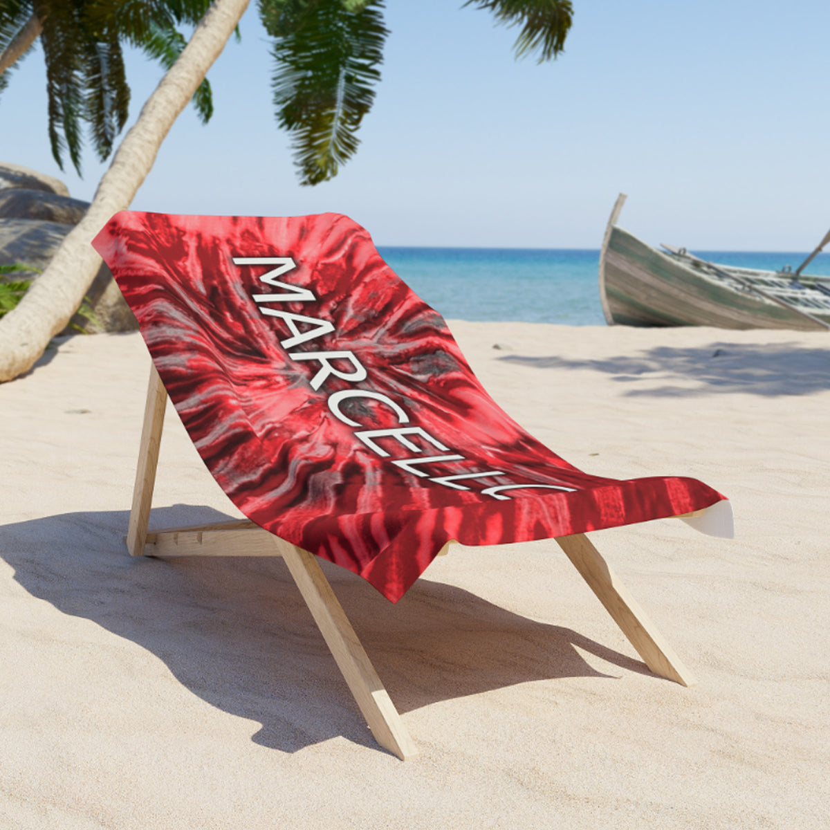 A red and black tie-dye beach towel with the name 'MARCELLE' printed in white, draped over a wooden beach chair on a sandy tropical beach.