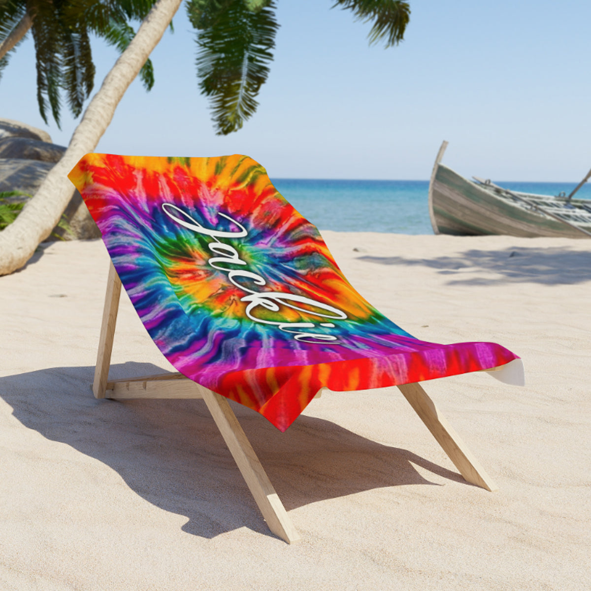 A colorful tie-dye beach towel draped over a wooden beach chair on a sandy beach, with palm trees, ocean, and a boat in the background. The towel features a rainbow spiral pattern with the words 'Jazz & Love' written in white script.