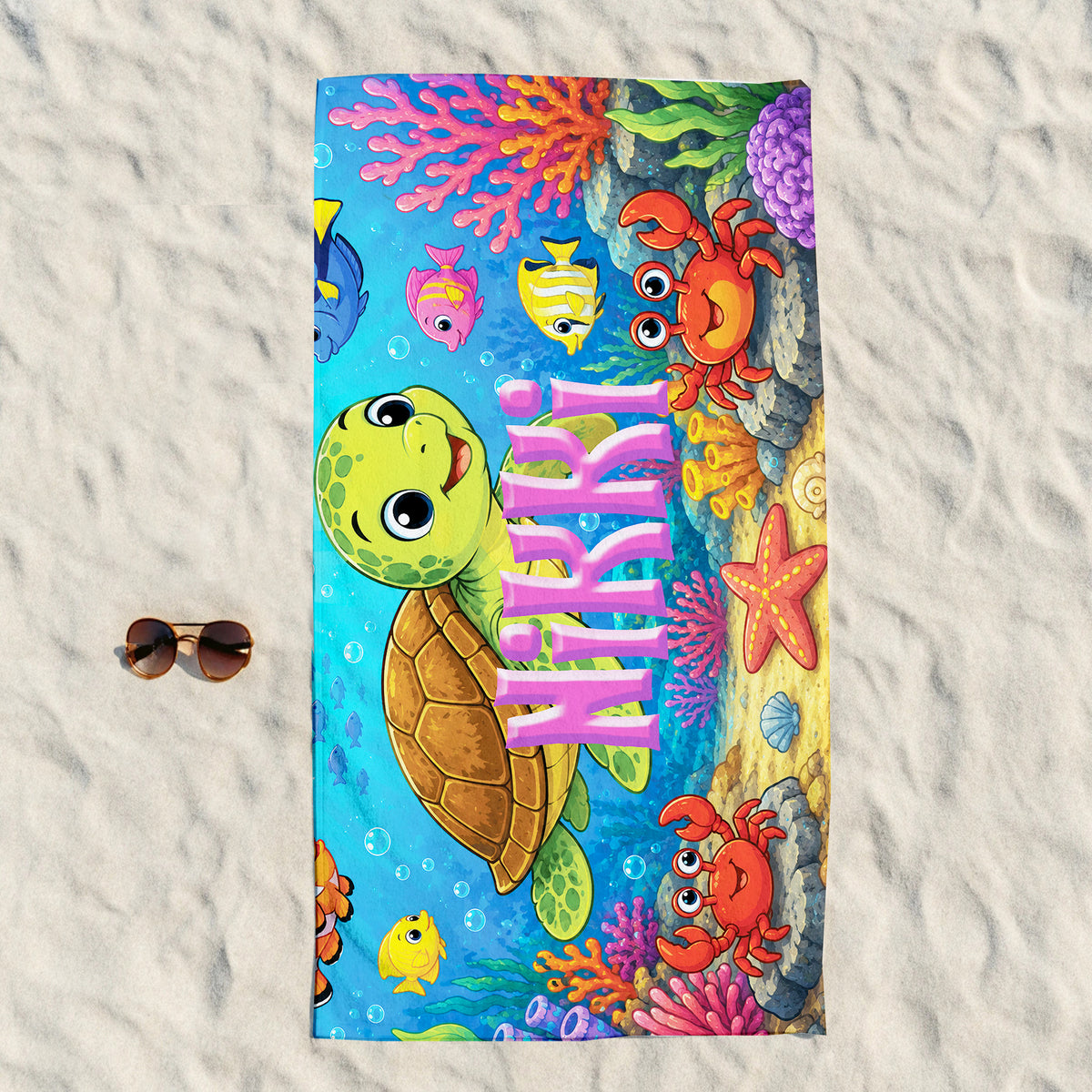 A colorful beach towel laid flat on white sand, featuring a cartoon sea turtle, crabs, fish, coral, and the word 'MERRY' in large pink letters. A pair of sunglasses rests to the left of the towel.