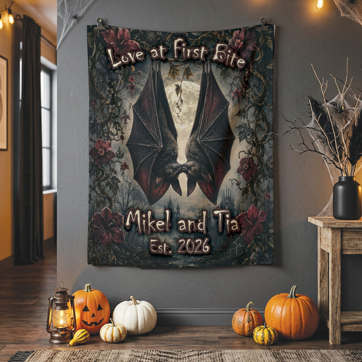 A Halloween-themed wall tapestry featuring a large bat silhouette, gothic floral border, and the text 'Love at First Bite' and 'Mikel and Tia Est. 2026', displayed on a gray wall in a decorated home interior with pumpkins, a lantern, and a side table.