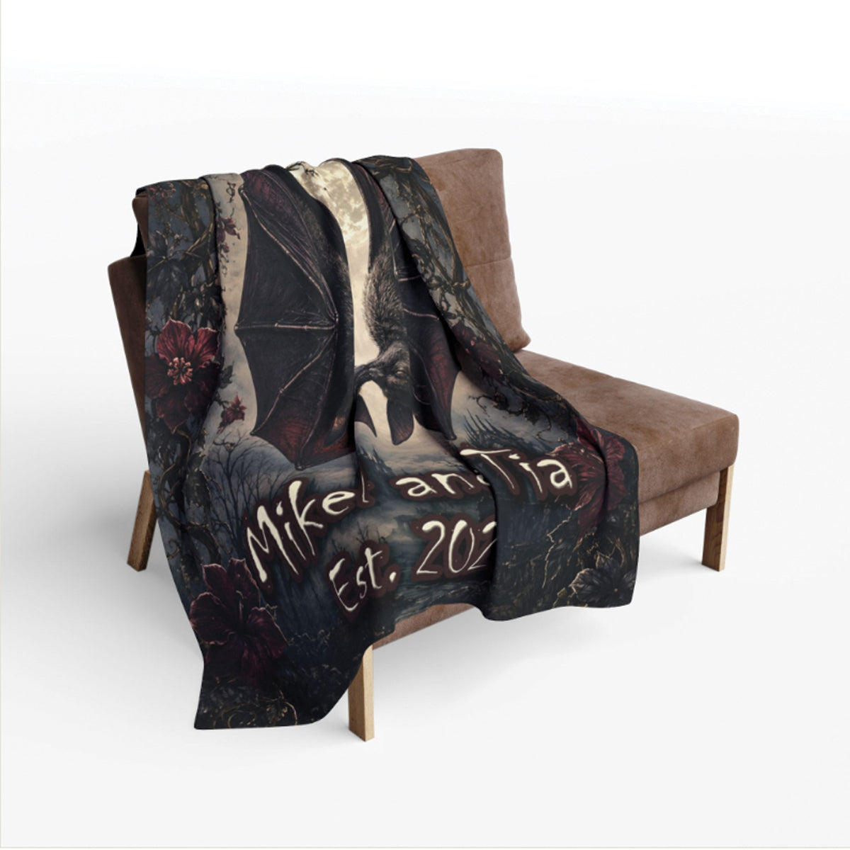 A dark printed throw blanket draped over a brown armchair against a light gray studio background. The blanket features a gothic illustration with bat wings, floral elements, and the text 'Mikaela Est.