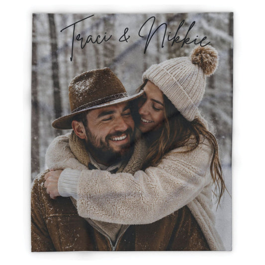 A square custom photo print featuring a couple embracing in the snow, with the names 'Traci & Nikki' written in black script at the top. The print is shown against a clean white background.