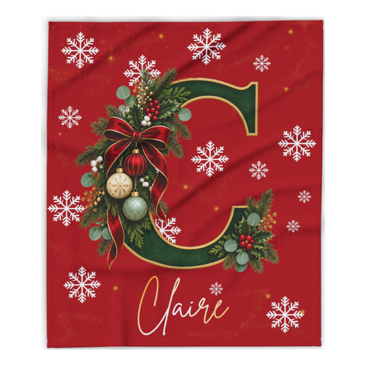 A red holiday-themed personalized blanket or fabric print featuring a large green letter C decorated with a red bow, green foliage, ornaments, and white snowflakes, with the name 'Claire' in white script at the bottom.