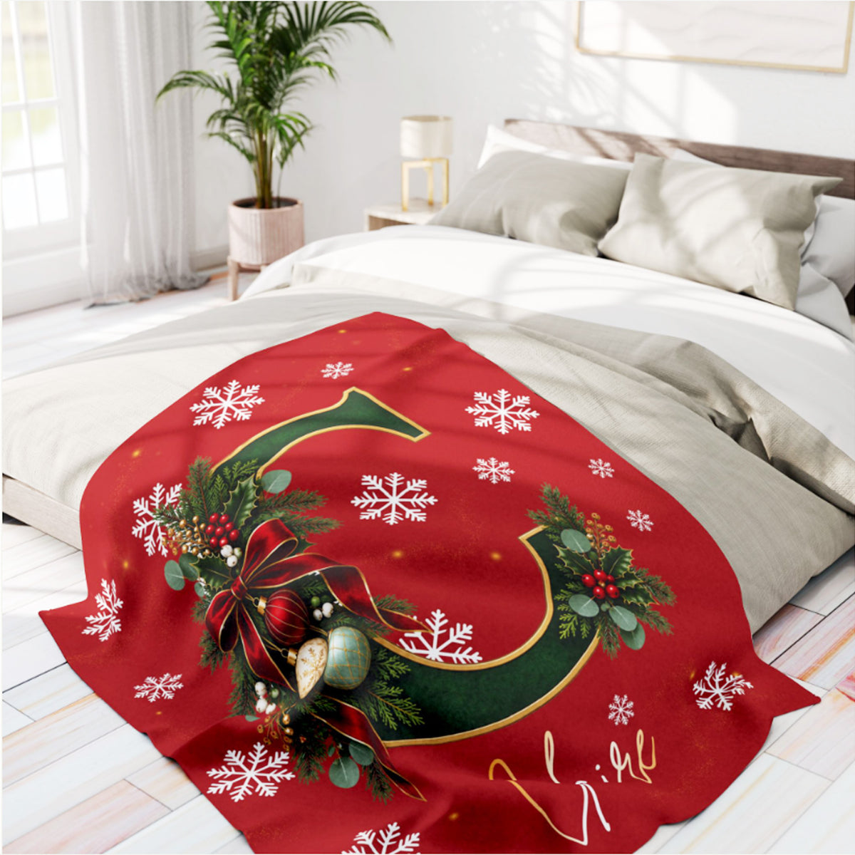 A festive red holiday blanket draped over a bed in a bright, modern bedroom. The blanket features a large green letter 'G', white snowflakes, green pine garlands with red berries, and a red bow.